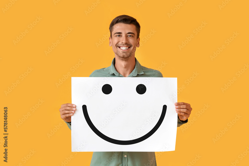 Smiling young man holding paper with happy emoticon on yellow background Smiling young man holding paper with happy emoticon on yellow background