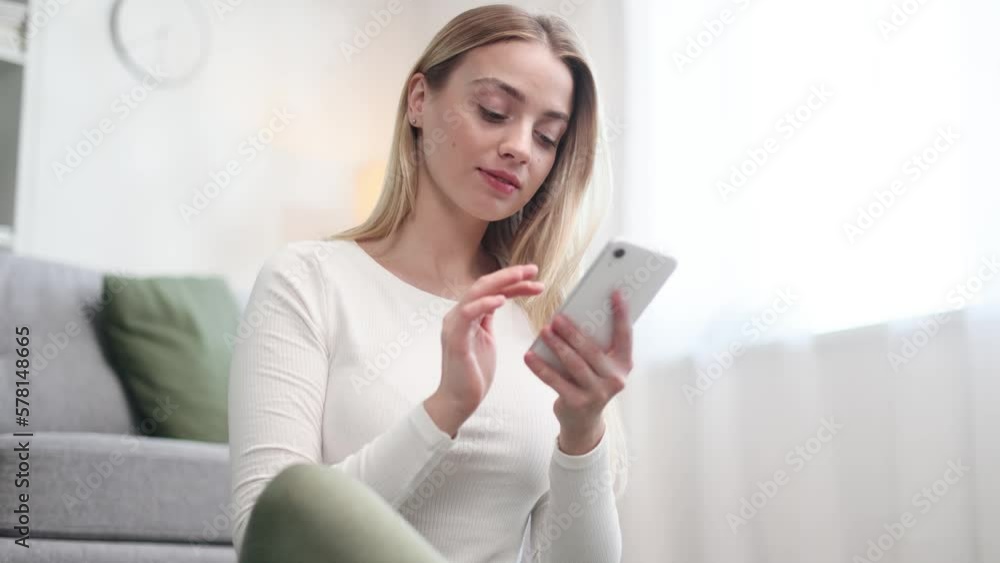 Attractive young blond woman using smartphone scrolling social media texting browsing online in the light living room Beautiful girl relaxing while chatting at mobile phone at home Attractive young blond woman using smartphone scrolling social media texting browsing online in the light living room Beautiful girl relaxing while chatting at mobile phone at home