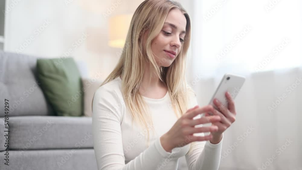 Portrait of beautiful blond woman hold smartphone watching social media with sport bottle take a break after training and looking at camera at home Smiling young woman texting on phone at light room Portrait of beautiful blond woman hold smartphone watching social media with sport bottle take a break after training and looking at camera at home Smiling young woman texting on phone at light room