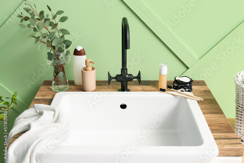 Table with ceramic sink, bath accessories and eucalyptus in vase near green wall
