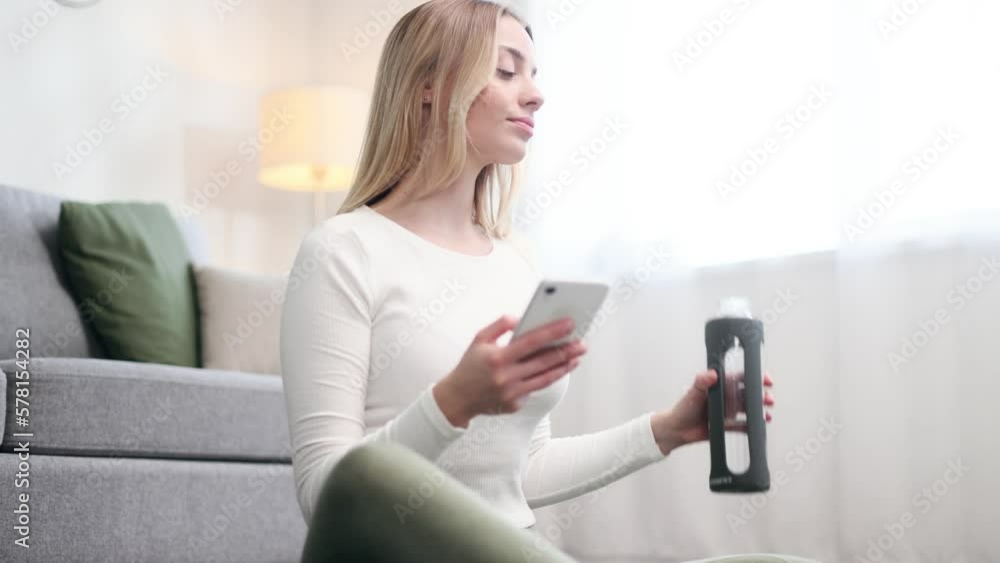 Portrait of beautiful blond woman hold smartphone watching social media drinking from sport bottle take a break after training at home Smiling young woman texting on her phone at light room Portrait of beautiful blond woman hold smartphone watching social media drinking from sport bottle take a break after training at home Smiling young woman texting on her phone at light room