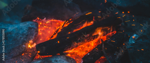 Vivid smoldered firewoods burned in fire close-up. Atmospheric background with orange flame of campfire. Unimaginable full frame image of bonfire. Glowing embers in air. Warm logs, bright sparks bokeh