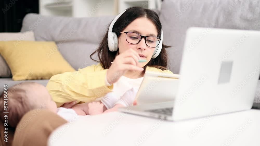 Portrait of busy young business mother holding her baby while working or studying on laptop computer and make notes at light home office Successful woman balancing work or education and motherhood Portrait of busy young business mother holding her baby while working or studying on laptop computer and make notes at light home office Successful woman balancing work or education and motherhood