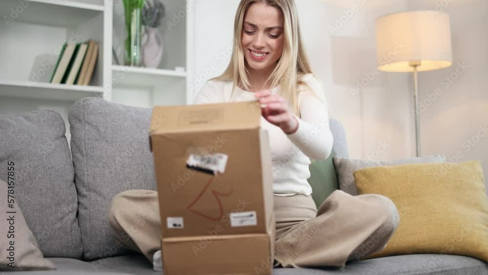 Portrait of blond young woman received parcel dissatisfied with shopping in online internet store sitting on sofa at home Upset customer disappointed with purchase order while opening carton box Portrait of blond young woman received parcel dissatisfied with shopping in online internet store sitting on sofa at home Upset customer disappointed with purchase order while opening carton box
