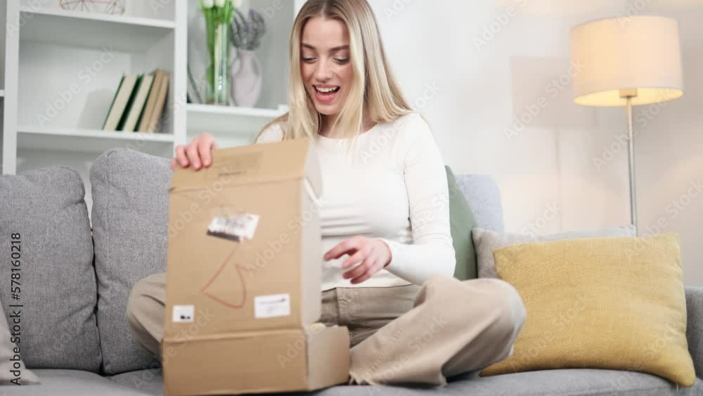 Charming young blond woman received parcel satisfied with shopping in online internet store sitting on sofa at home Happy customer rejoice with purchase order while opening carton box indoors Charming young blond woman received parcel satisfied with shopping in online internet store sitting on sofa at home Happy customer rejoice with purchase order while opening carton box indoors