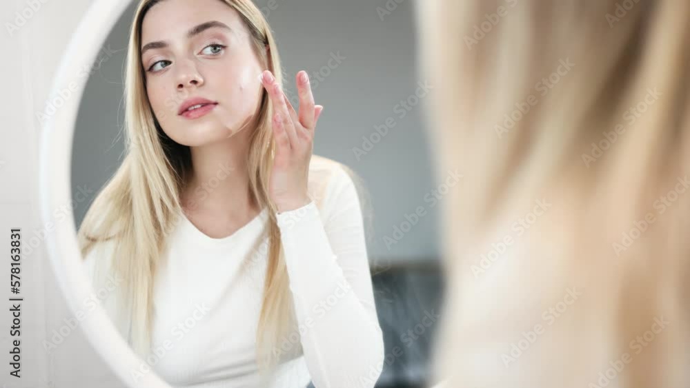 Portrait of pretty young blond woman looking at mirror reflection and applying face cream moisturising skin at home Concept of skincare cosmetology natural beauty cosmetics Portrait of pretty young blond woman looking at mirror reflection and applying face cream moisturising skin at home Concept of skincare cosmetology natural beauty cosmetics