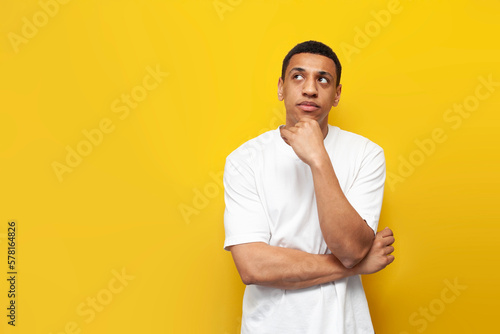 young pensive afro american guy in white oversized t-shirt stands on yellow isolated background and looks to the side