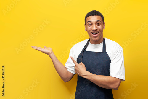 young guy afro american waiter in apron advertises copy space on yellow isolated background, barista worker