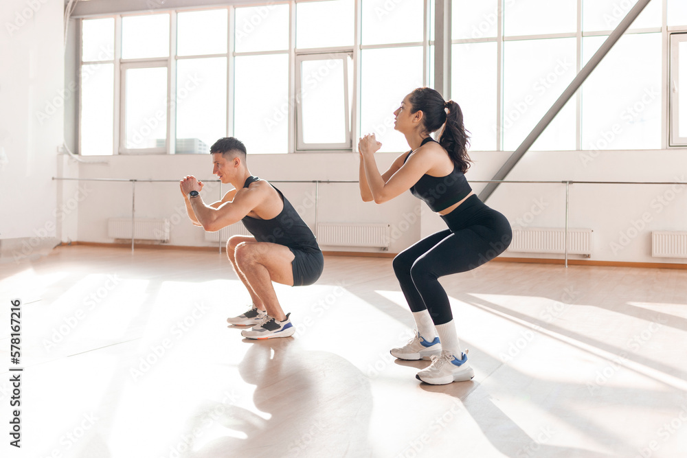 Fototapeta premium athletic couple in sportswear squat in the gym in the morning, fitness girl and trainer man train in light fitness room