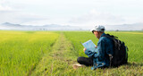Asian boy in plaid shirt wears cap and has a backpack, holding a map and a binoculars, sitting on ridge rice field of asian farmers to study landscapes, mountains, rice paddy field, bird watching.