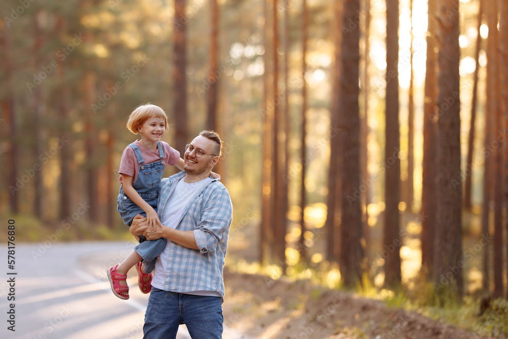 Fototapeta premium A magical moment unfolds as a father holds little daughter in his arms and they laugh joyously while strolling along a forest road, surrounded by majestic pine trees and the warm glow of the sunset.