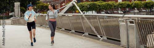 Two athletic women in sportswear is jogging on a bridge and listening music. Reaching the goal
