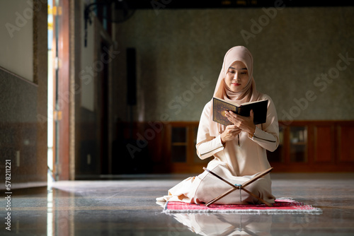 The image of an Asian Muslim woman in the Islamic religion in hijab in cream color. Sitting reading the Quran and having a happy smiling face Staying in a beautiful mosque out of respect for God.