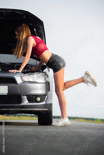 Beautiful young girl stands near the broken car with open hood on the empty road. © Dmitriy