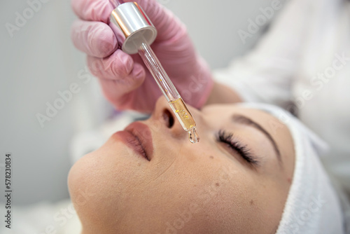 Cosmetologist applying few drops of moisturizing oil with a pipette on face her young pretty woman
