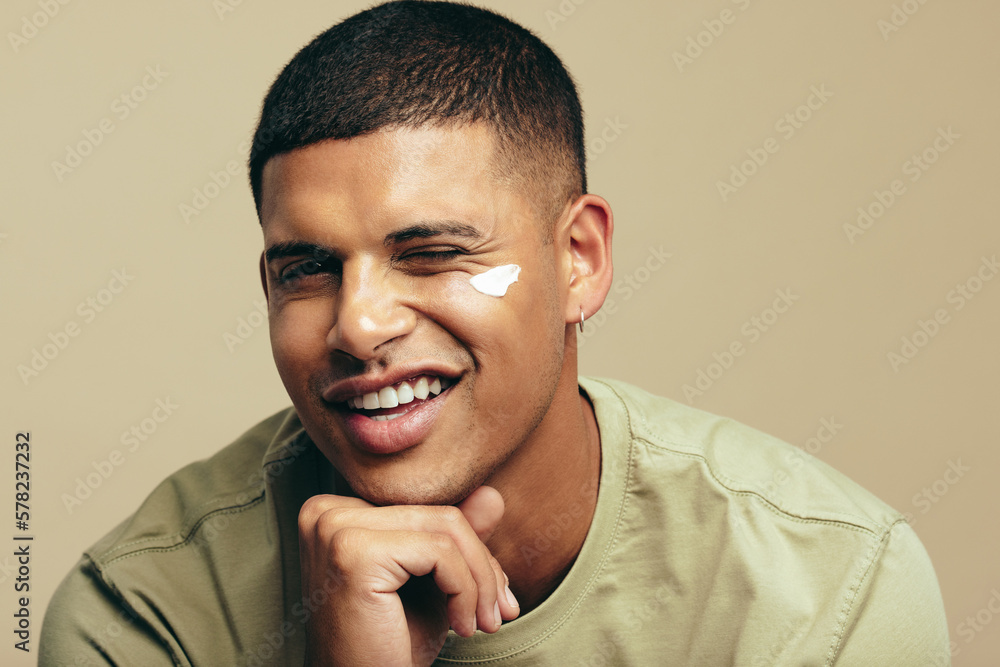 Obraz premium Fun skincare routine: Handsome young man smiles with beauty cream on his face