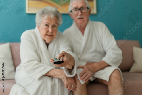 Happy senior couple sitting at sofa in bathrobes and watching TV.