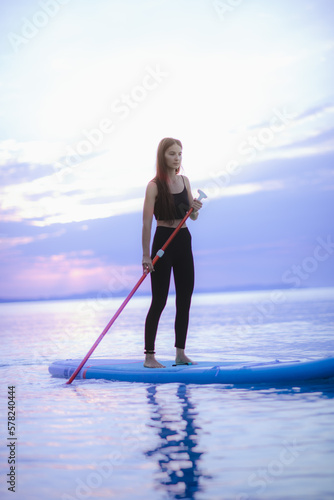 Young beautiful girl surfer paddling on surfboard on the lake at sunrise