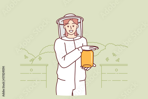 Young woman beekeeper in protective uniform holding jar of honey standing in apiary among flying bees. Girl farmer breeds bees in garden getting organic honey for sale at fair of ecological products