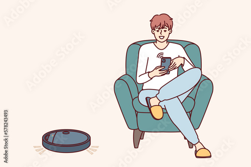 Robotic vacuum cleaner guided using mobile phone cleans floor in apartment near man sitting in chair. Robot vacuum cleaner with wi-fi chip is controlled via IOT and integrated into smart home system