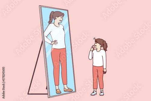 Little girl stands and looks in mirror, saw reflection of adult woman and dreams of becoming adult as soon as possible. Preteen girl wants to be as beautiful as mother or older sister