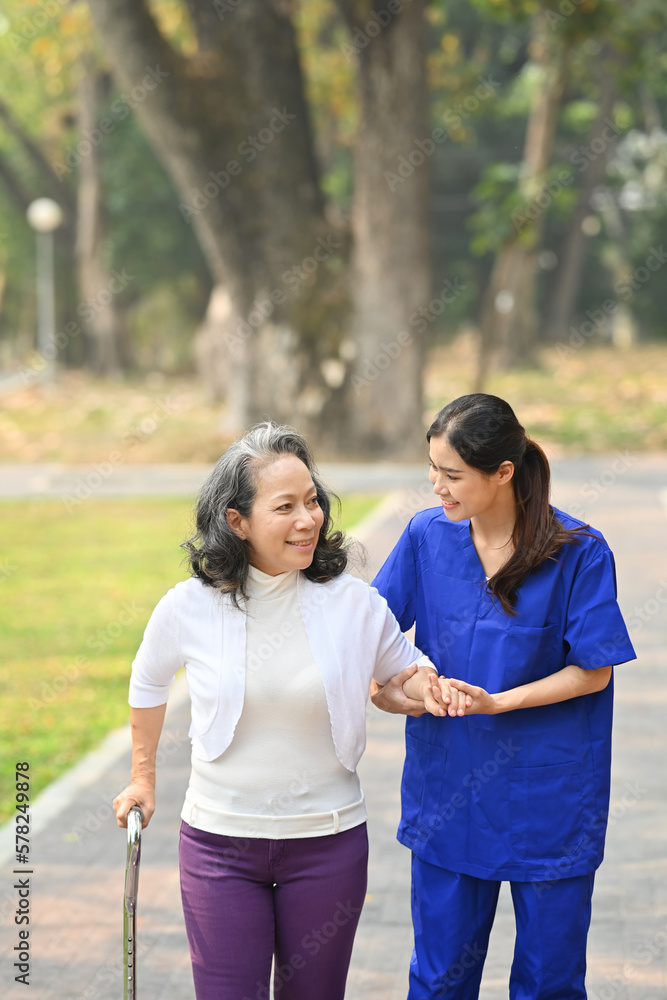 Image of caring female caregiver talking, supporting, helping elderly woman while walking in outdoor Image of caring female caregiver talking, supporting, helping elderly woman while walking in outdoor