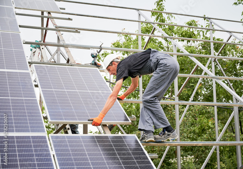 Worker installing solar panels on metal beams. Man in helmet and uniform. Photo-voltaic collection of modules. Array as a system of photo-voltaic panels. Concept of renewable and ecological energy. © anatoliy_gleb