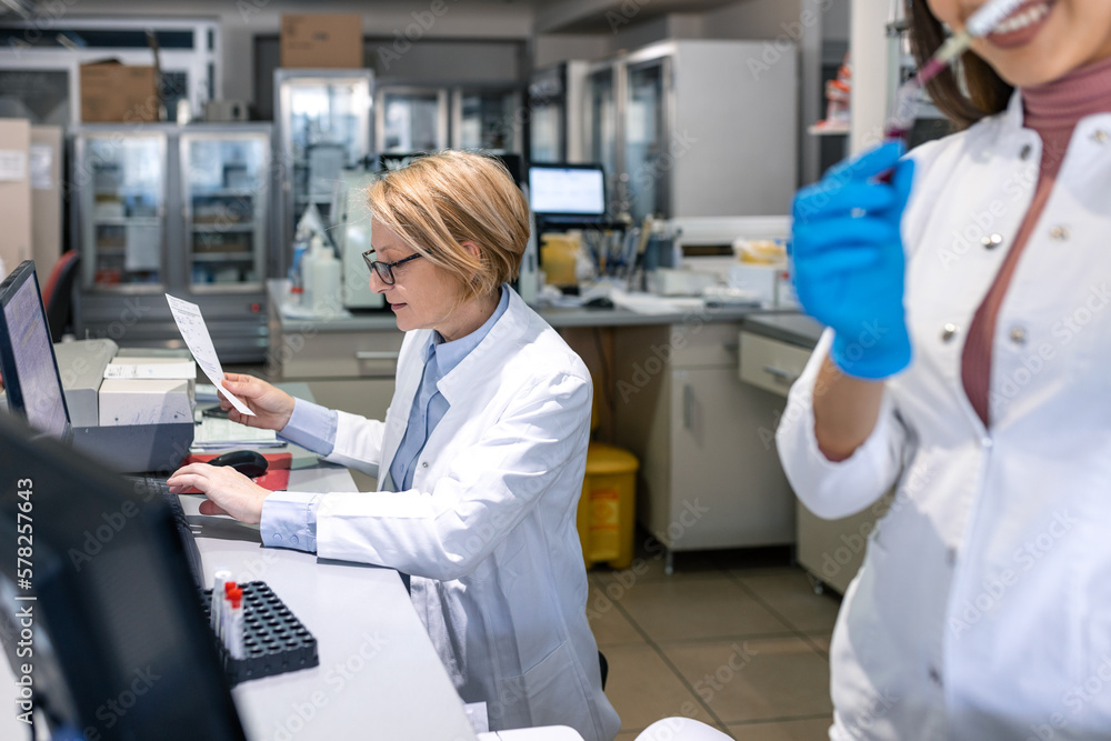 Female Scientists Working on their Computers In Big Modern Laboratory. Various Shelves with Beakers, Chemicals and Different Technical Equipment is Visible. Female Scientists Working on their Computers In Big Modern Laboratory. Various Shelves with Beakers, Chemicals and Different Technical Equipment is Visible.