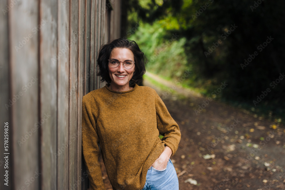 Happy woman with hand in pocket leaning on wooden wall Happy woman with hand in pocket leaning on wooden wall