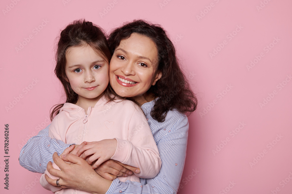 Multi-ethnic pretty woman, loving mother smiles a cheerful toothy smile looking at camera, experiencing happiness while gently hugging her adorable beloved little child girl, isolated pink background