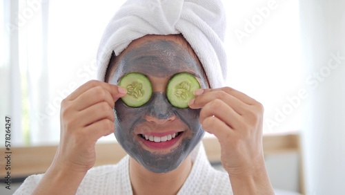 Asian woman with grey mask on face is putting slices of cucumber on eyes smiling and look at camera. Spa beauty procedures at home for girl. Skin Care, hydrating and moisturising concept