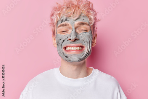 Portrait of cheerful adult man with broad toothy smile keeps eyes closed enjoys skin care beauty procedures applies facial clay mask dressed in casual white t shirt isolated over pink background. photo