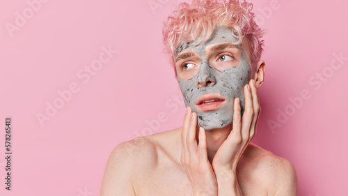 Horizontal shot of thoughtful man keeps hands on cheeks focused aside attentively applies facial nourishing clay mask stands shirtless against pink background copy space for your advetising content photo