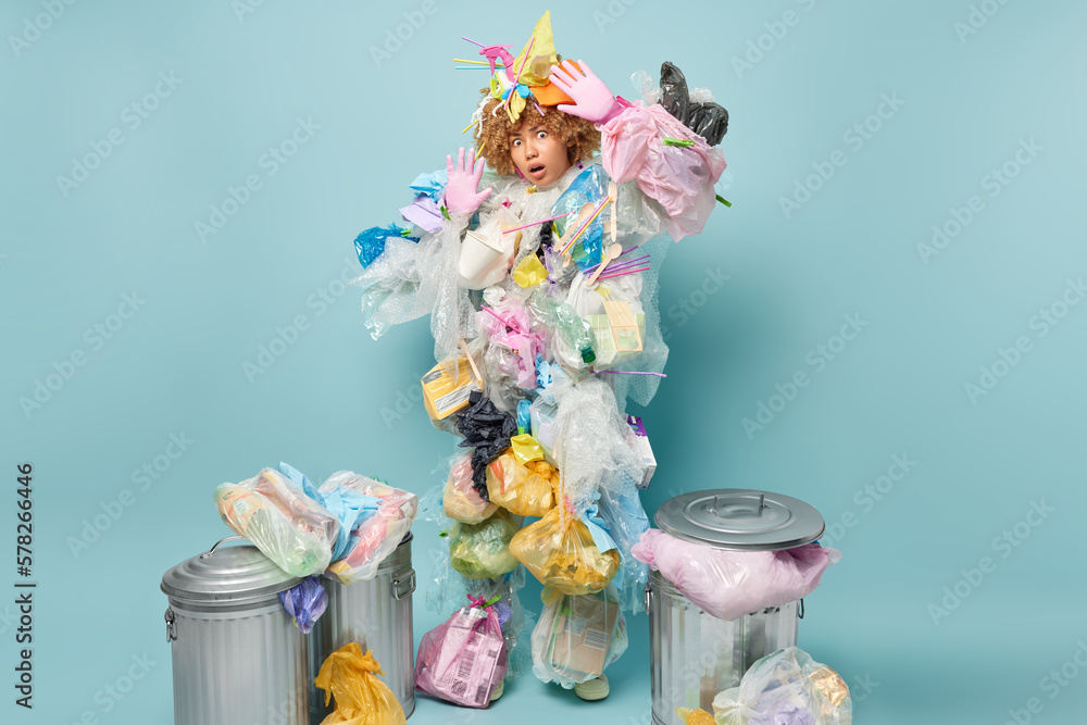 Scared shocked curly haired woman keeps palms raised tries to protect herself from danger wears plastic garbage costume stares impressed at camera surrounded by litter bins busy picking up wastes Scared shocked curly haired woman keeps palms raised tries to protect herself from danger wears plastic garbage costume stares impressed at camera surrounded by litter bins busy picking up wastes