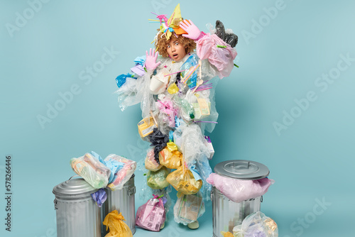 Scared shocked curly haired woman keeps palms raised tries to protect herself from danger wears plastic garbage costume stares impressed at camera surrounded by litter bins busy picking up wastes