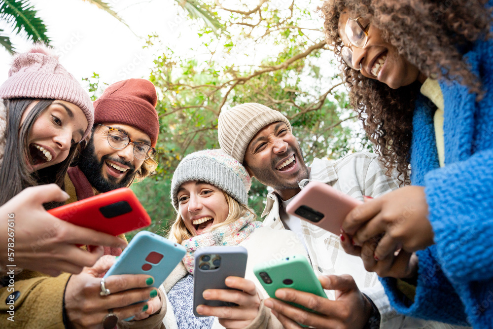 Obraz premium Group of happy multi ethnic friends sharing social media content on smart phone, winter outdoors