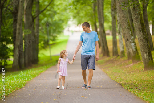 Little daughter and young adult father speaking and walking on sidewalk through tree alley at city park. Spending time together in beautiful warm sunny summer day. Front view. © fotoduets