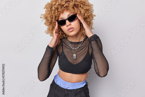 Indoor shot of stylish curly haired young woman wears trendy sunglasses black net top and trousers poses in fashionable clothes against white studio background going to have walk during sunny day.