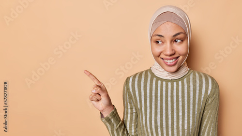 Photography Horizontal shot of happy Muslim woman points index finger left shows promo offer