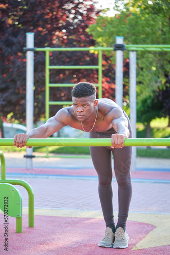 Young fit shirtless black man doing push-ups on a bar in a calisthenics park outdoors on sunny day. Fitness and sport lifestyle.
