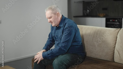 An elderly man is sitting on the couch at home and is experiencing pain in his knee. A sick joint causes a lot of inconvenience to a pensioner