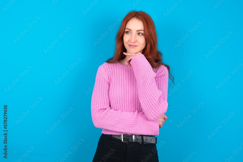 Dreamy young woman wearing pink sweater over blue background with pleasant expression, looks sideways, keeps hand under chin, thinks about something pleasant. Dreamy young woman wearing pink sweater over blue background with pleasant expression, looks sideways, keeps hand under chin, thinks about something pleasant.