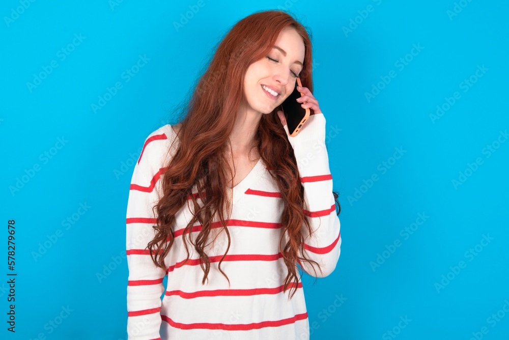 Funny young woman wearing striped T-shirt over blue background laughs happily, has phone conversation, being amused by friend, closes eyes. Funny young woman wearing striped T-shirt over blue background laughs happily, has phone conversation, being amused by friend, closes eyes.