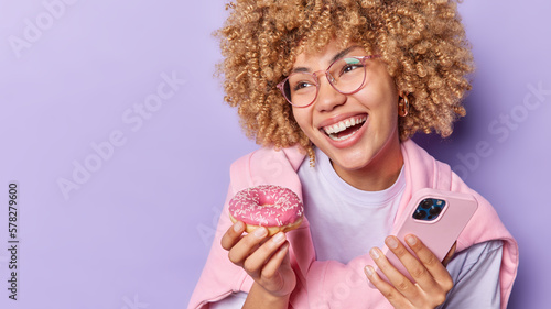 Horizontal shot of cheerful curly woman wears transparent eyeglasses and pullover holds delicious appetizing doughnut and smartphone eats appetizing snack isolated over purple background blank space