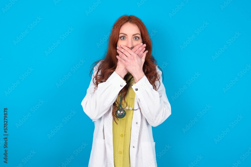 Stunned young doctor woman wearing medical uniform over blue background covers both hands on mouth, afraids of something astonishing Stunned young doctor woman wearing medical uniform over blue background covers both hands on mouth, afraids of something astonishing