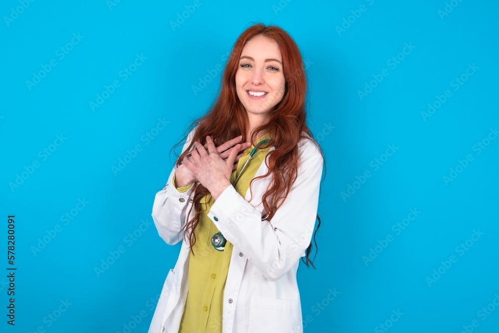 young doctor woman wearing medical uniform over blue background expresses happiness, laughs pleasantly, keeps hands on heart young doctor woman wearing medical uniform over blue background expresses happiness, laughs pleasantly, keeps hands on heart
