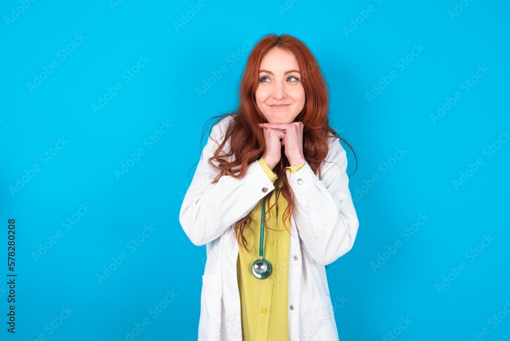 Curious young doctor woman wearing medical uniform over blue background keeps hands under chin bites lips and looks with interest aside. Curious young doctor woman wearing medical uniform over blue background keeps hands under chin bites lips and looks with interest aside.
