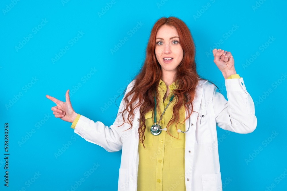 young doctor woman wearing medical uniform over blue background points at empty space holding fist up, winner gesture. young doctor woman wearing medical uniform over blue background points at empty space holding fist up, winner gesture.