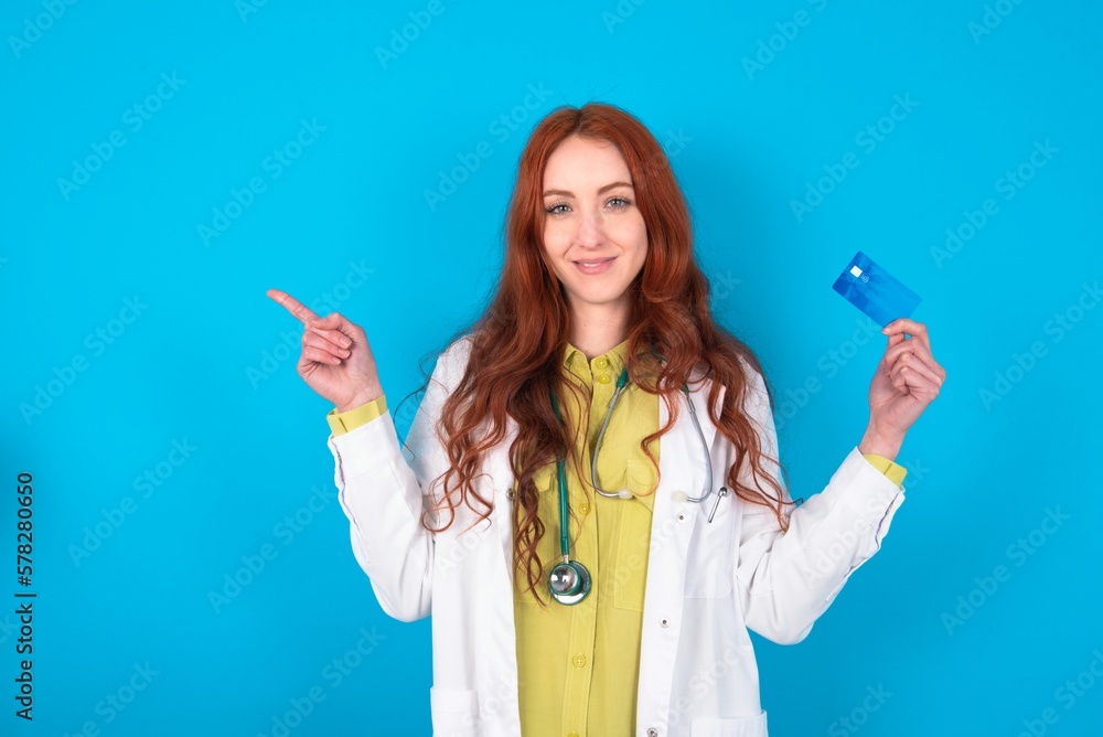 Smiling young doctor woman wearing medical uniform over blue background showing debit card pointing finger empty space Smiling young doctor woman wearing medical uniform over blue background showing debit card pointing finger empty space