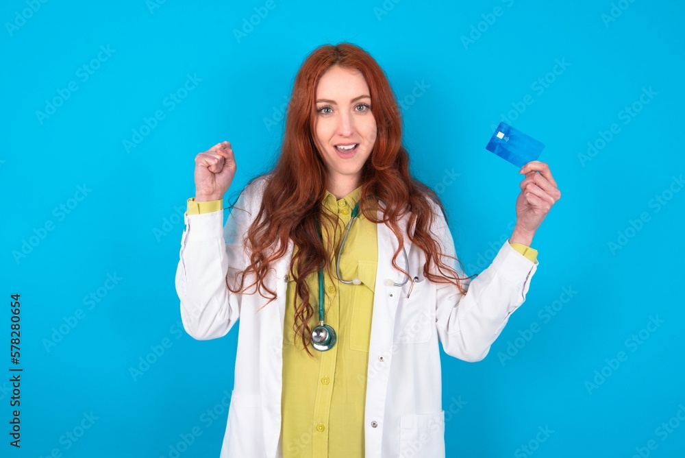 Photo of lucky impressed young doctor woman wearing medical uniform over blue background arm fist holding credit card. Celebrated Photo of lucky impressed young doctor woman wearing medical uniform over blue background arm fist holding credit card. Celebrated